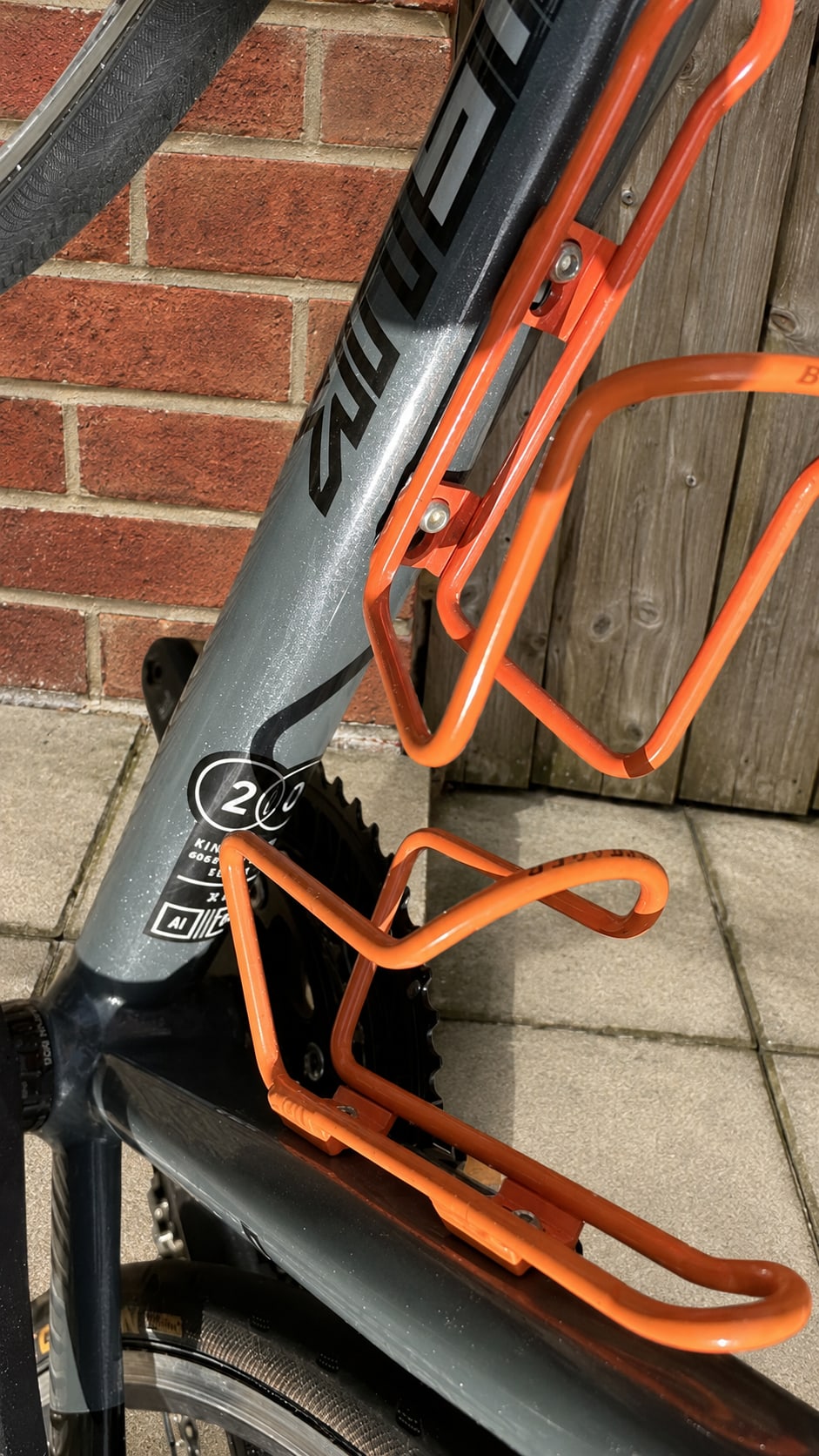 Orange and black bike frame detail showing seat tube and bottle cage mounts against brick wall and wooden decking, close-up angle.