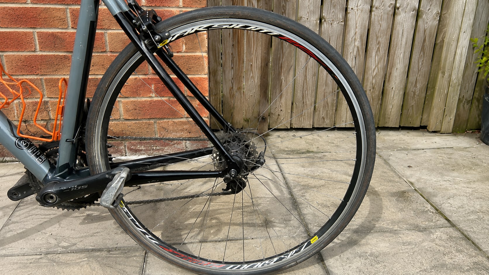 Rear wheel of a grey and black road bike with disc brakes, non-drive side, against a brick wall and wooden fence.