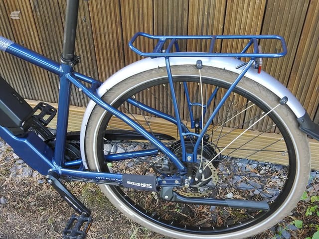 Blue Nottingham Hybrid HESC utility bike with white mudguards and blue rear rack, drive side, on ground against corrugated metal backdrop.