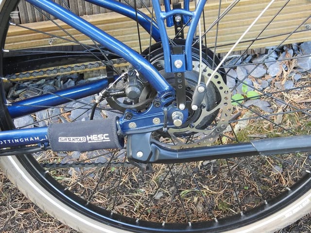 Blue hybrid bike rear brake and drivetrain detail, drive side, on ground with mulch and wooden fence background.