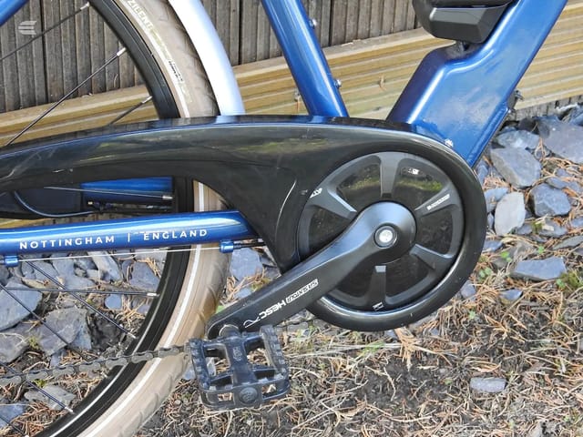 Blue Nottingham England bike, drive side, close-up of crankset and pedal on gravel ground with wooden decking and fence behind.