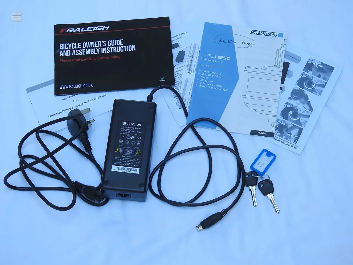 Electric bicycle documentation and accessories laid flat on a light blue background, including Raleigh owner's guide, Suntour manual, Phylion battery charger,