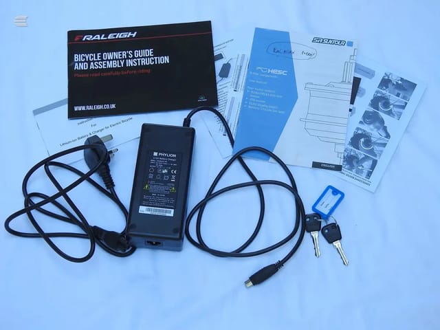 Electric bicycle documentation and accessories laid flat on a light blue background: Raleigh owner's guide, Suntour manual, Phylion battery charger with