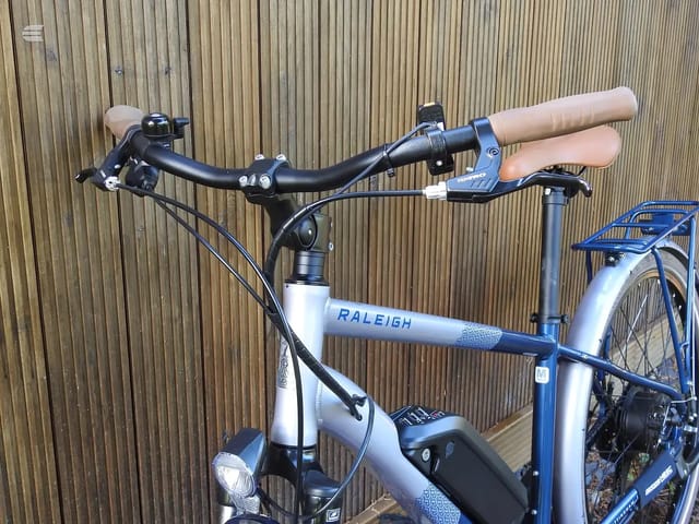 Blue and white Raleigh hybrid bike, non-drive side, against corrugated metal shutter. Handlebars and saddle visible, front wheel cropped.