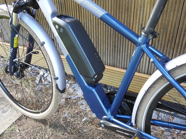Blue and white electric hybrid bike on wooden decking, three-quarter view from drive side, against corrugated metal backdrop. Battery mounted on down tube.