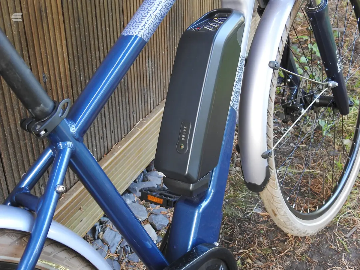 Blue and white electric hybrid bike on a wooden deck, three-quarter view, drive side, with battery pack visible on seat tube.