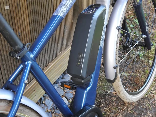 Blue and white electric hybrid bike on a wooden deck, three-quarter view, drive side, with battery pack visible on seat tube.