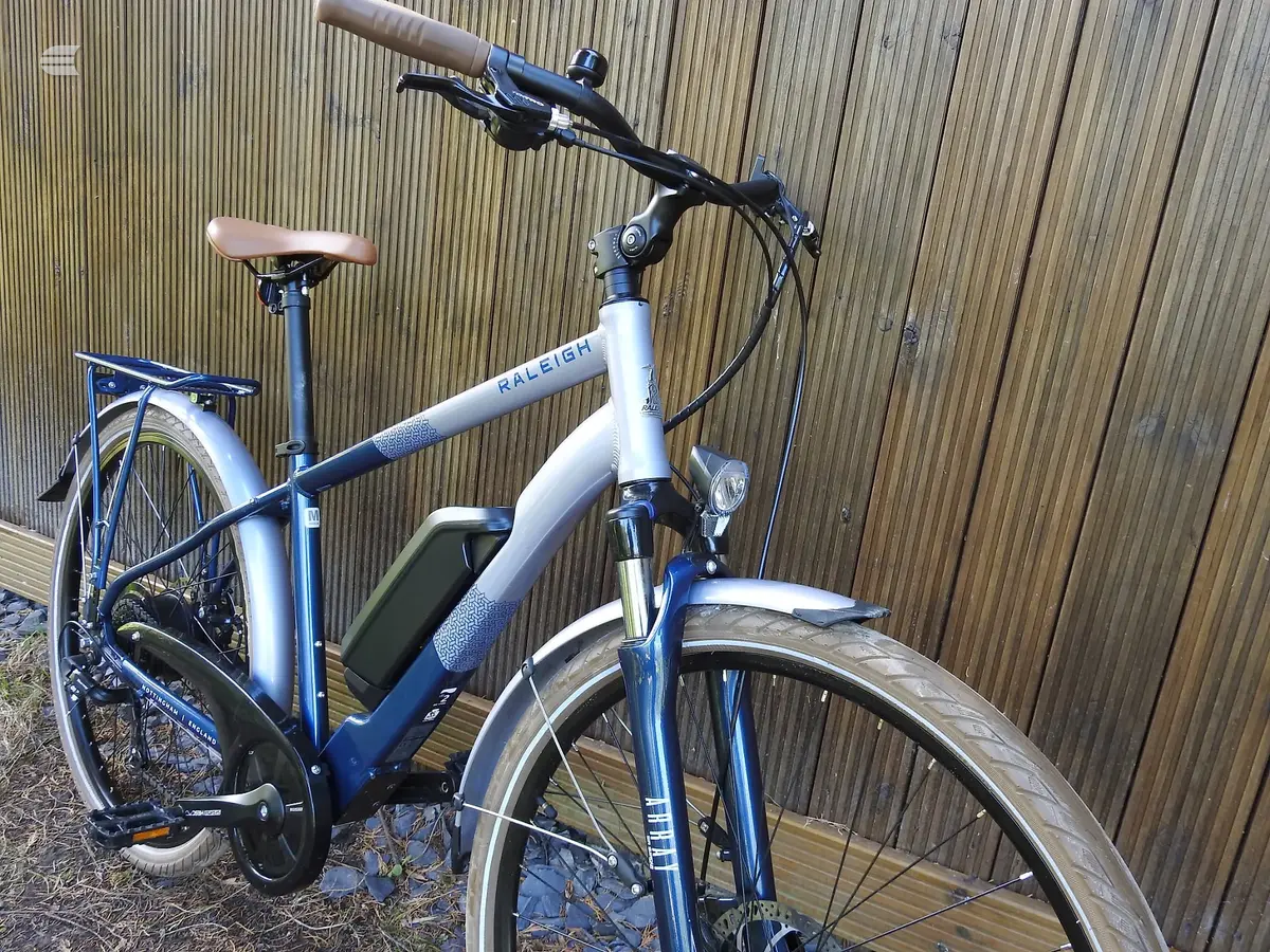 Blue and white Raleigh electric hybrid bike, three-quarter view from drive side, against corrugated metal shed. Mudguards and rear rack visible.