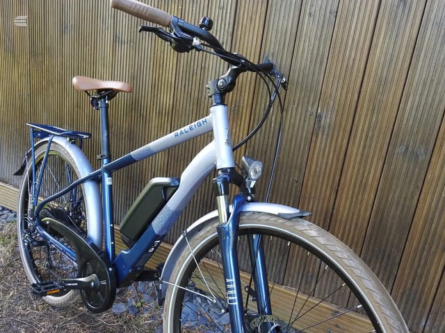 Blue and white Raleigh electric hybrid bike, three-quarter view from drive side, against corrugated metal shed. Mudguards and rear rack visible.