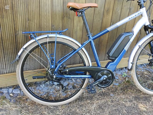 Blue and white Raleigh hybrid bike with brown saddle, drive side, parked on wooden decking against corrugated metal backdrop.