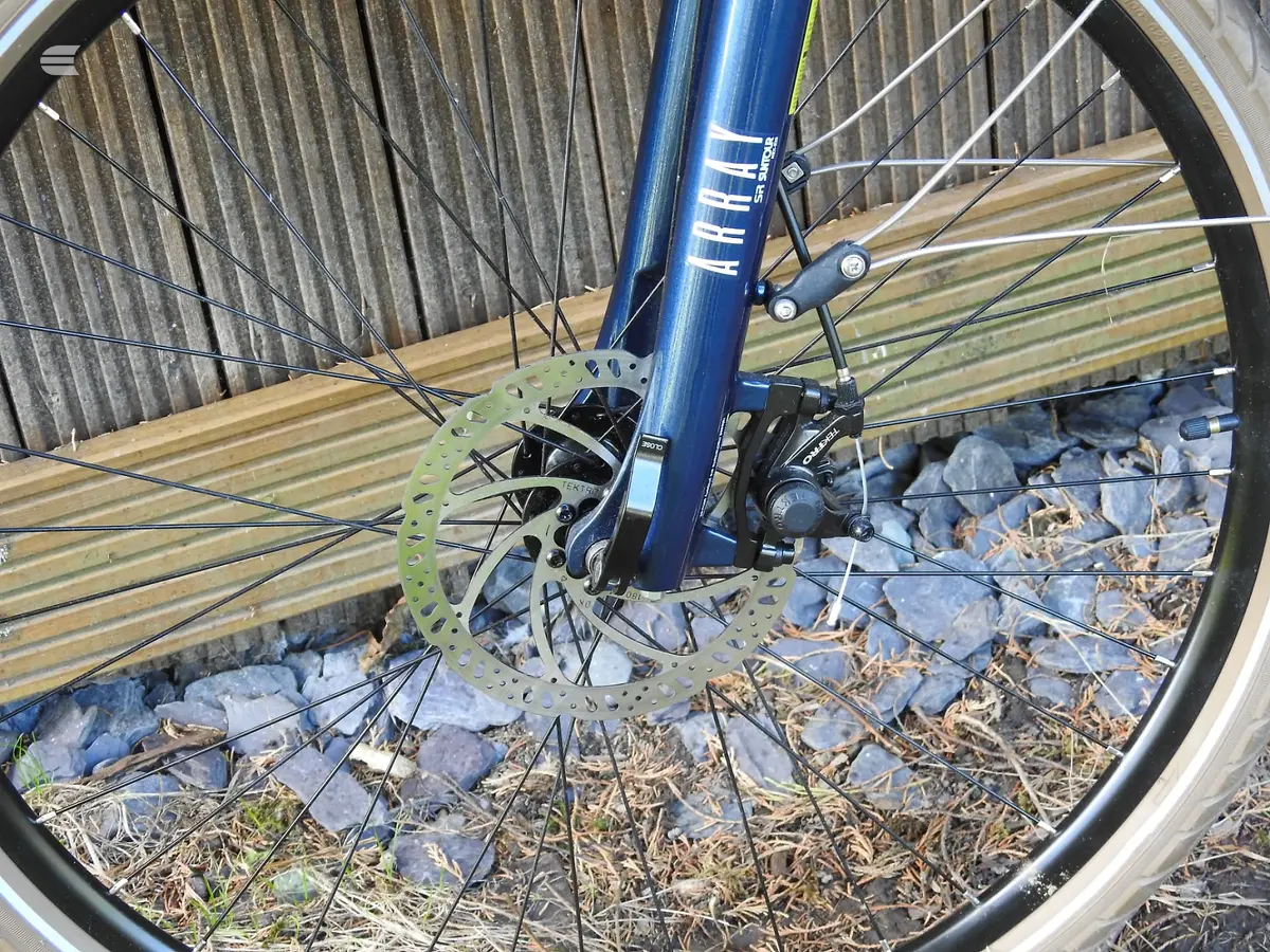 Blue hybrid bike front wheel with hydraulic disc brake and green rotor, drive side, on wooden decking and gravel.