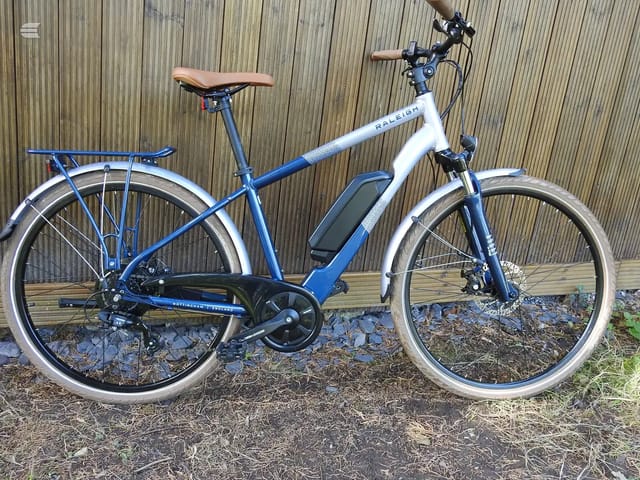 Blue and white Raleigh electric hybrid bike with brown saddle, drive side, parked against corrugated metal shed with mudguards and front rack visible.