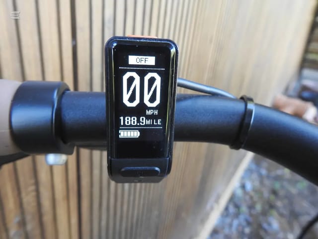 Black e-bike display console mounted on handlebars, showing 00 MPH, 188.9 mile odometer, and battery indicator, against corrugated metal background.