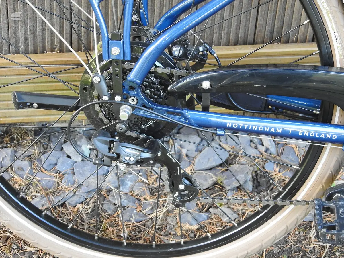 Blue Nottingham bicycle, rear derailleur and cassette close-up, drive side, on wooden decking with gravel background.