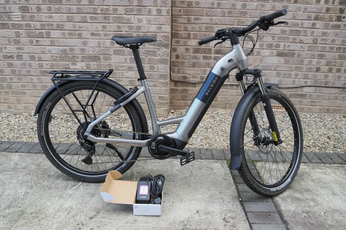 Silver and blue Raleigh electric hybrid bike with rear rack, parked on pavement against brick wall, non-drive side, with charger and accessories in cardboard