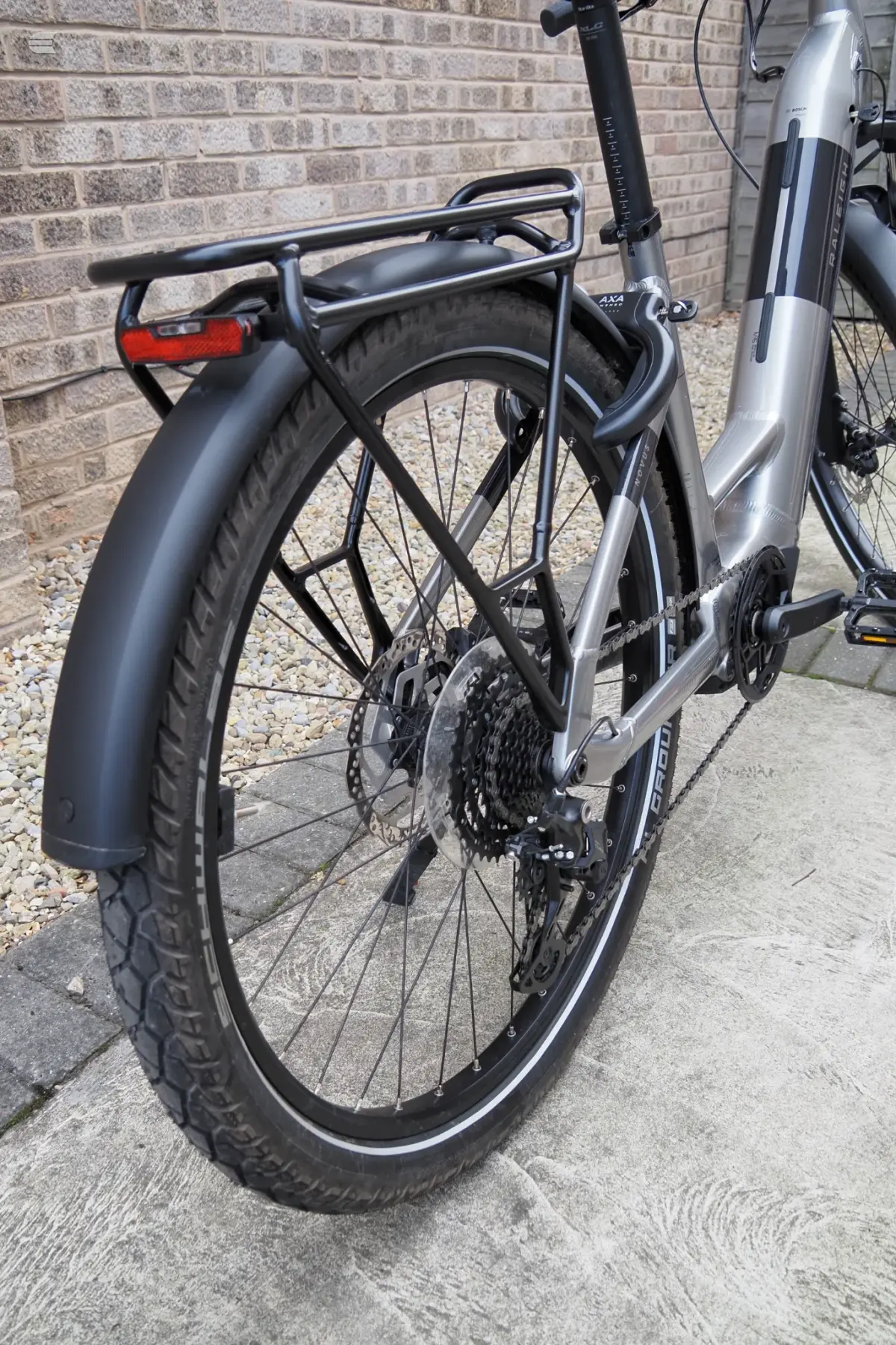 Silver and black electric cargo bike on a driveway, three-quarter drive-side view, against a brick wall. Features fat tyres, rear rack, and front suspension.