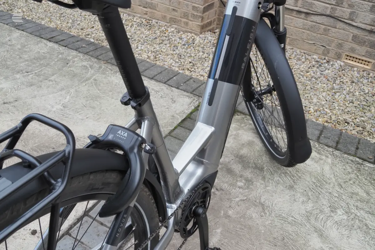 Grey and black Bosch e-bike with drop bars, parked on concrete driveway, non-drive side, against brick wall and gravel background.