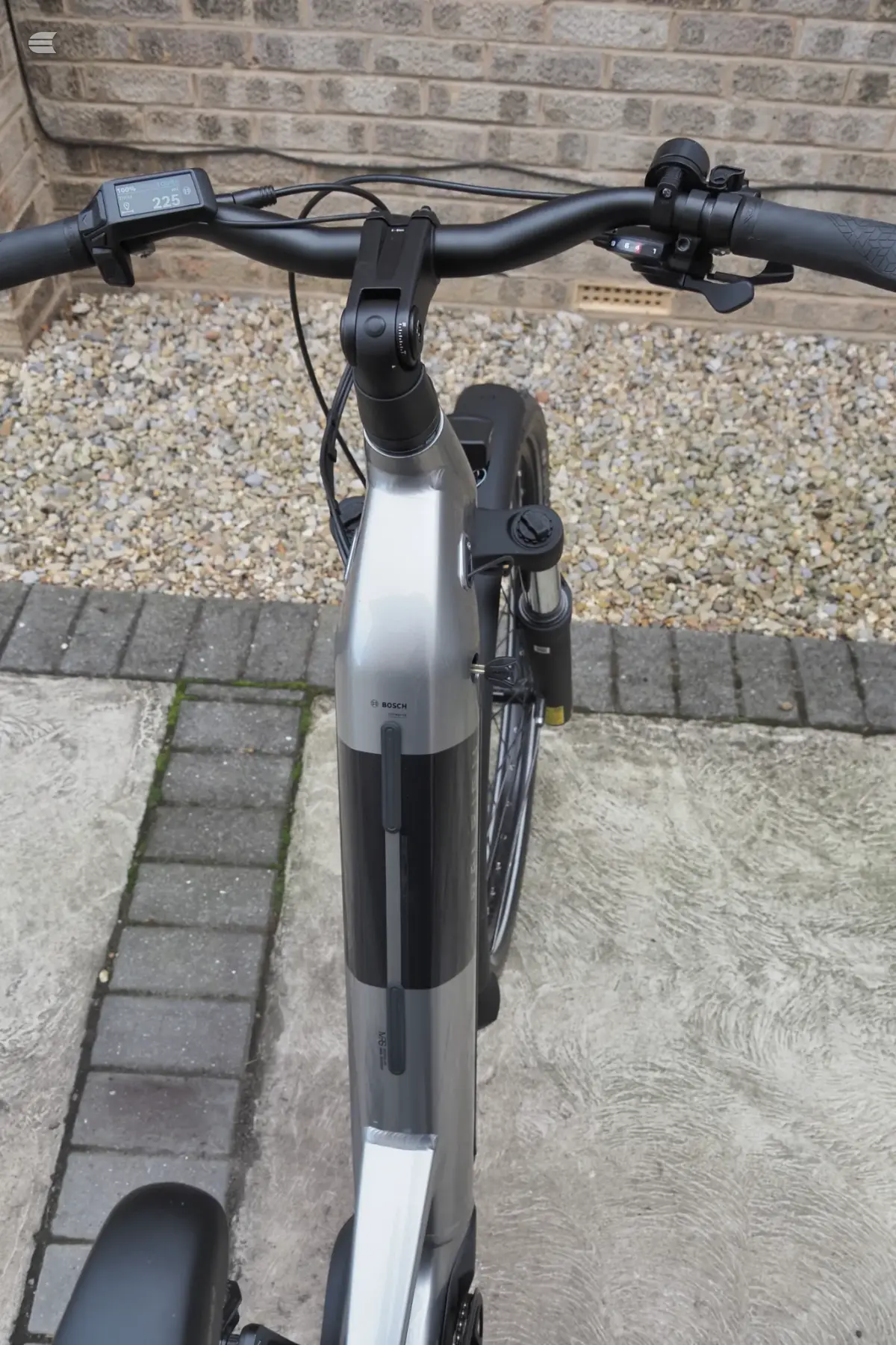 Silver and black Bosch e-bike, front-on view, parked on pavement against brick wall and gravel background.