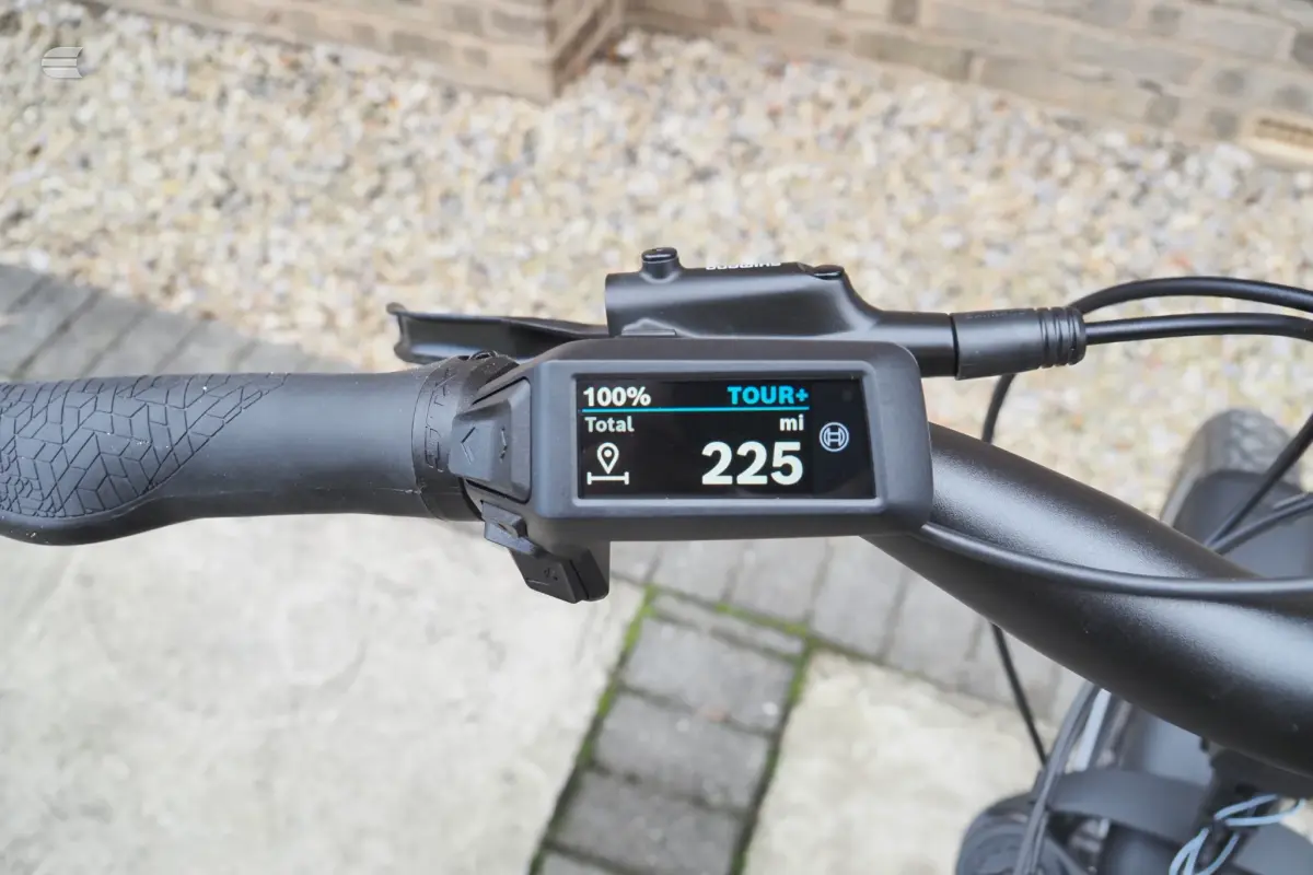 E-bike display console mounted on handlebar showing 225 miles total range on TOUR+ mode, against gravel background.