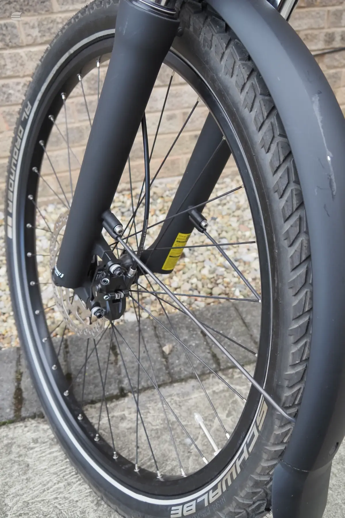 Front wheel of a dark grey gravel bike with disc brakes, drive side, against a brick wall and gravel ground.