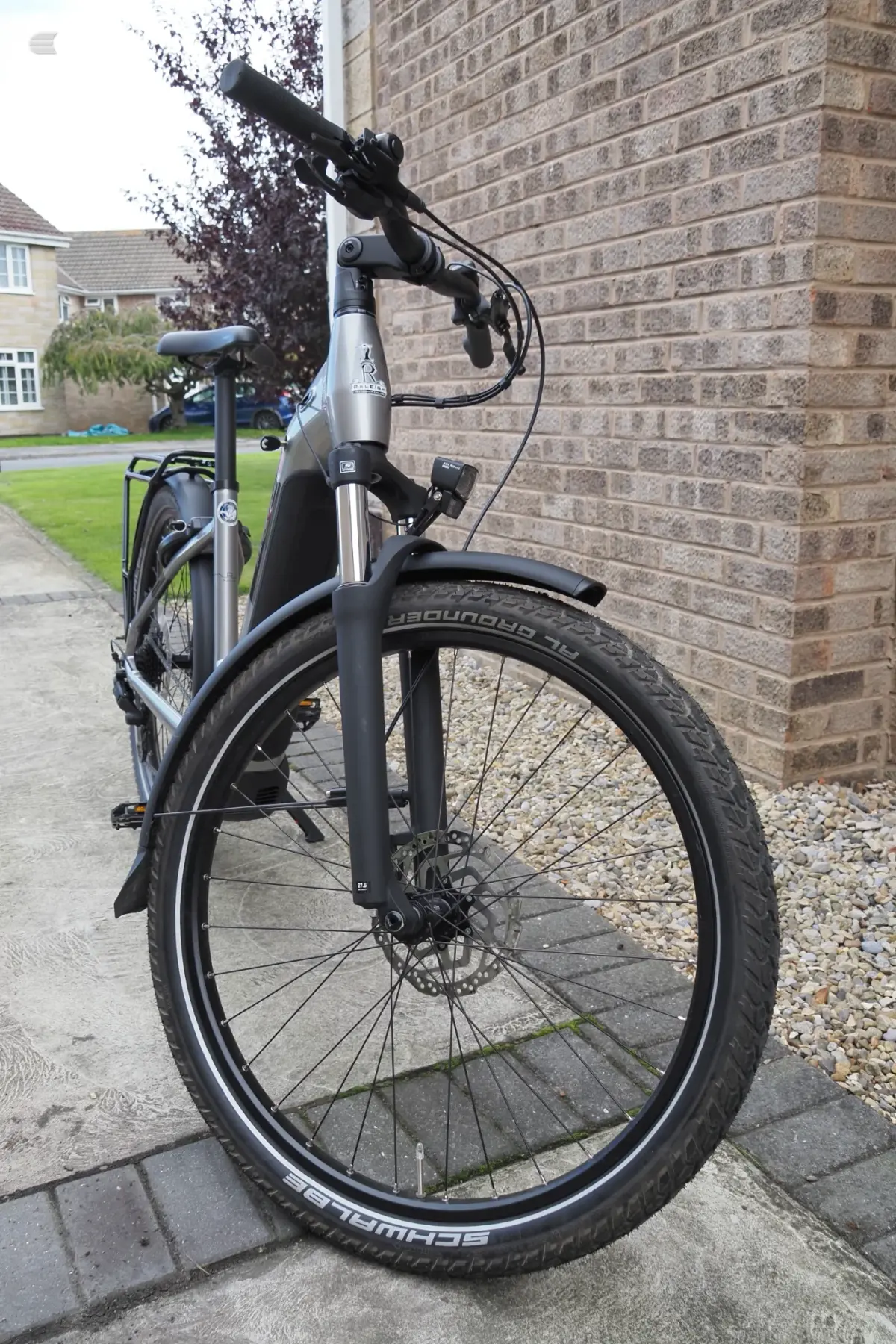 Silver and black hybrid electric bike on a driveway, non-drive side, leaning against a brick house wall.