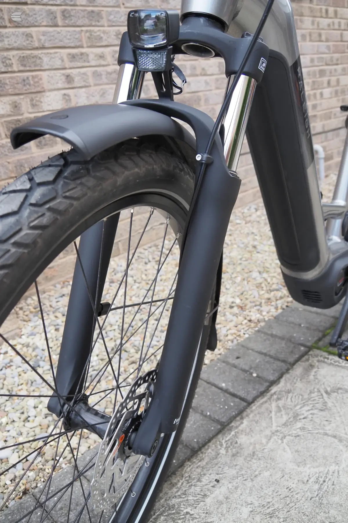 Black e-bike front wheel and fork close-up, non-drive side, against brick wall and gravel ground. Mudguard and front light visible.