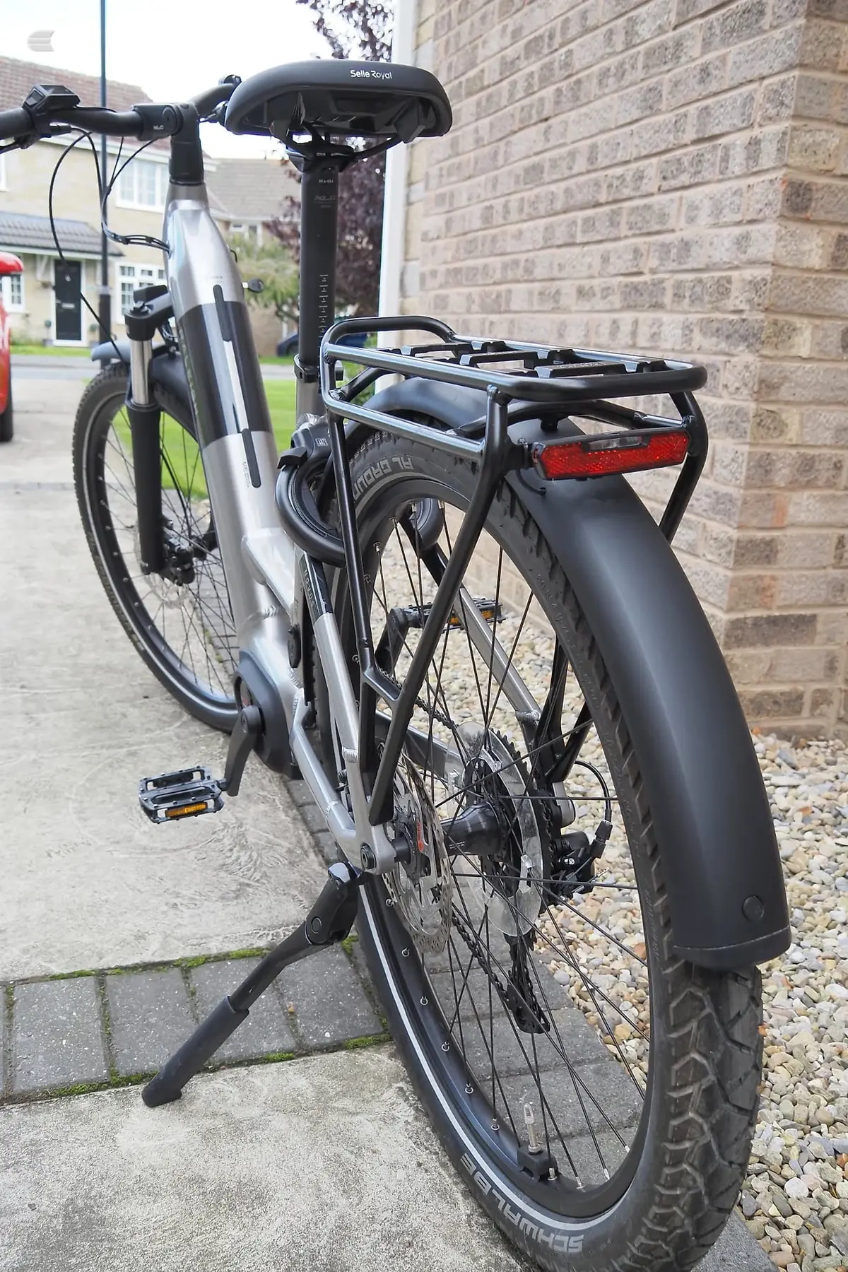 Silver and black electric bike on a stand, rear three-quarter view, parked against a brick wall on a residential driveway.