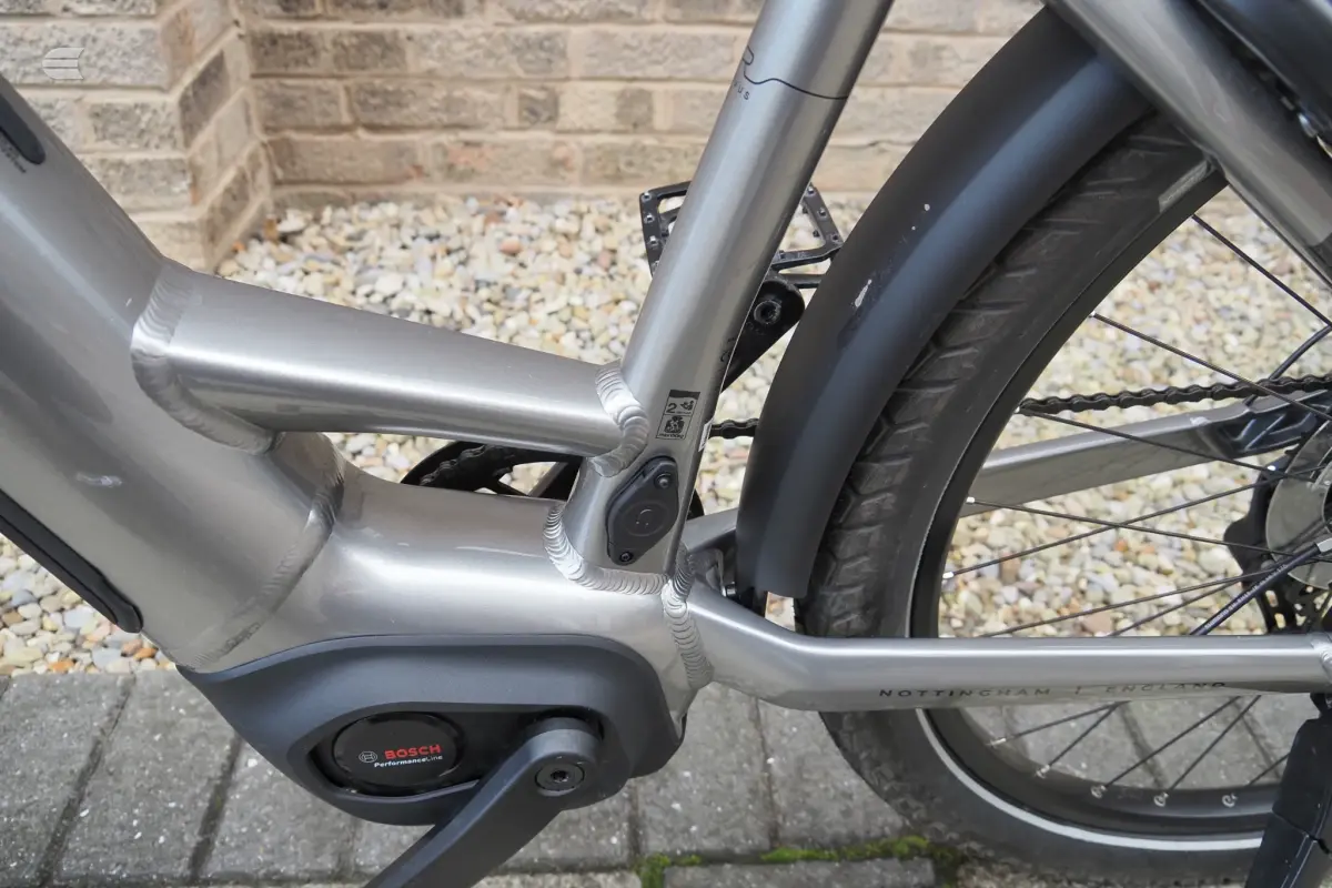 Silver Bosch mid-drive e-bike motor and frame detail, close-up of drive side, against brick wall and gravel.