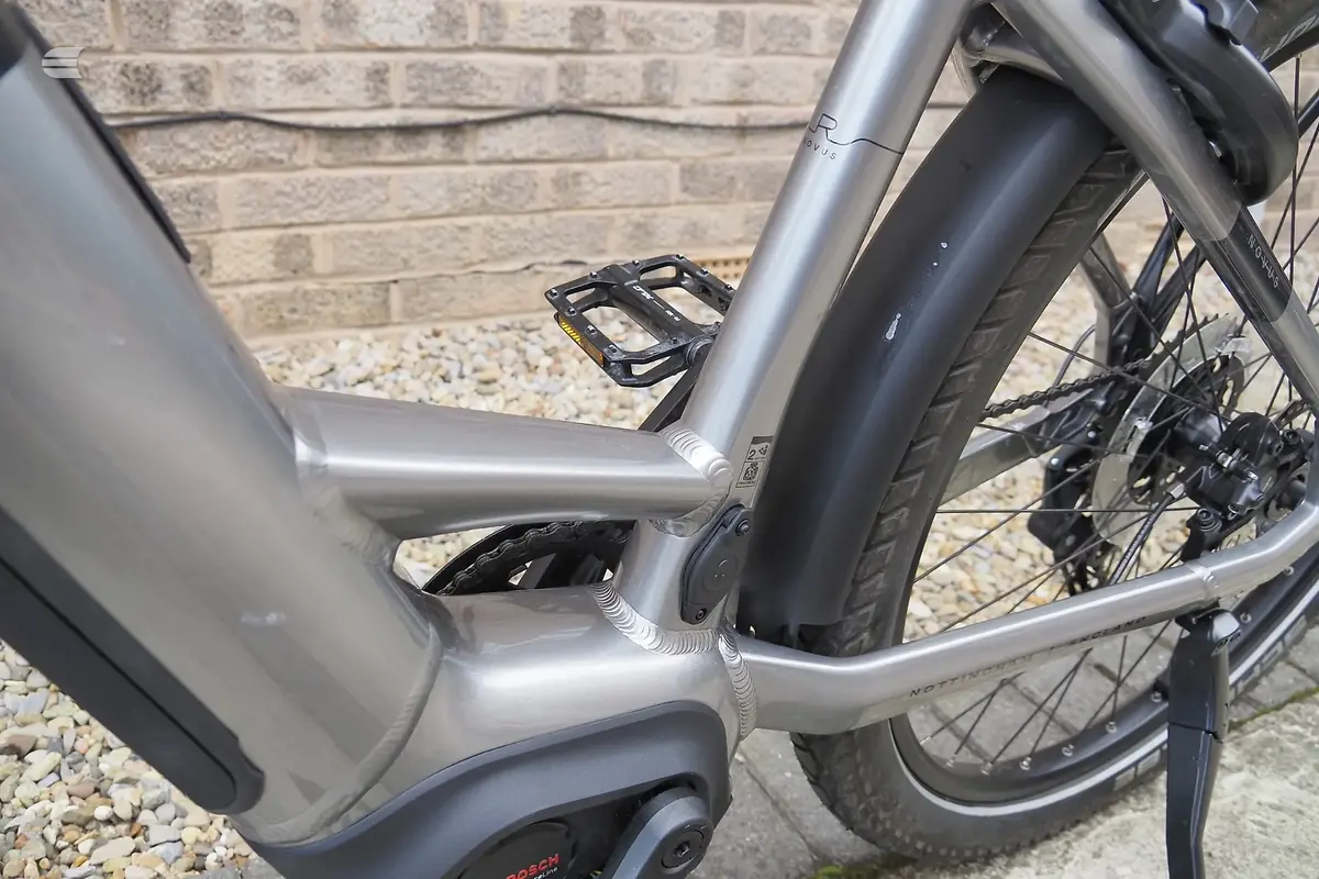 Silver and black e-bike frame detail, non-drive side, showing seat tube and down tube against brick wall and gravel.