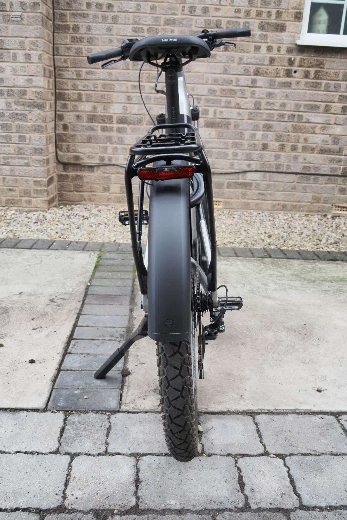 Black and grey electric cargo bike, rear three-quarter view, parked on paved driveway against brick house wall.
