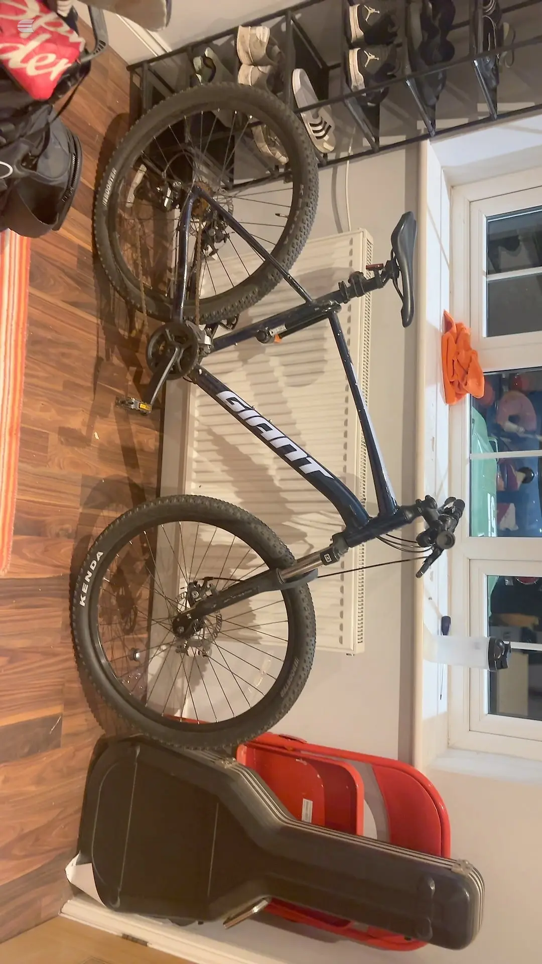 Black hardtail mountain bike mounted vertically on wall rack, drive side visible, in residential interior with white door and wooden flooring.