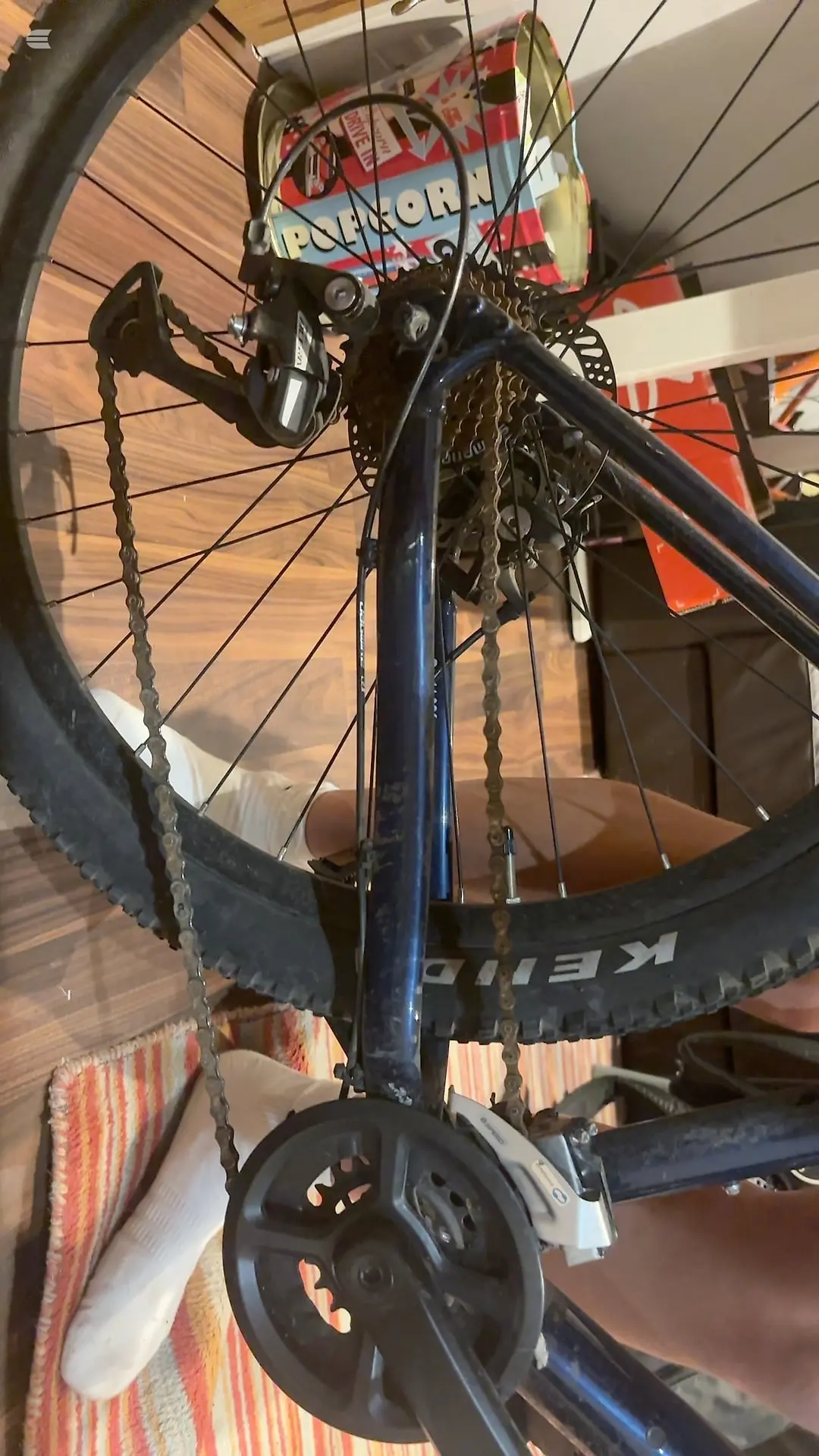 Blue mountain bike indoors on wooden floor, drive side, with red and white branded seat bag visible on rear rack.