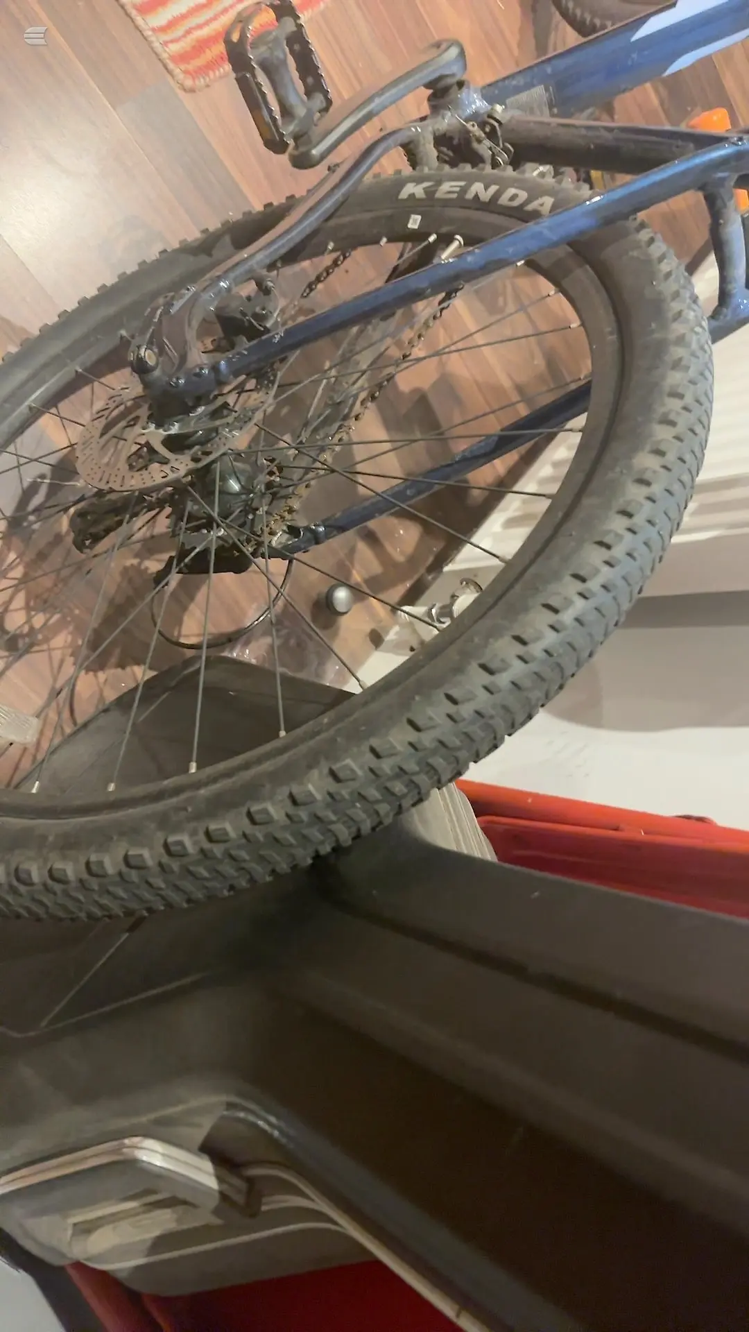 Blue mountain bike rear wheel and drivetrain, close-up angled view indoors on wooden floor, Kenda tyre visible.