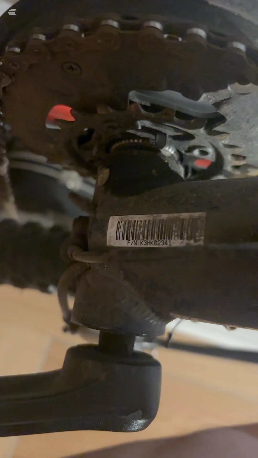 Close-up of bicycle brake caliper and internal components, dark metal parts with red accents, barcode label visible, on tan surface.