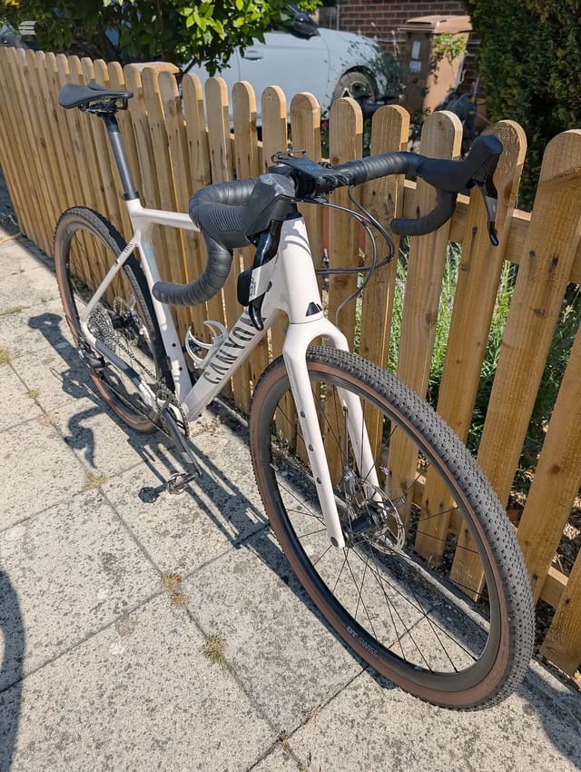 White and black gravel bike with drop bars, drive side, parked against wooden fence on paved ground in bright sunlight.
