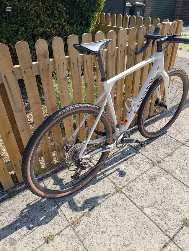 White gravel bike with black handlebars and saddle, drive side, leaning against wooden picket fence on paved ground.