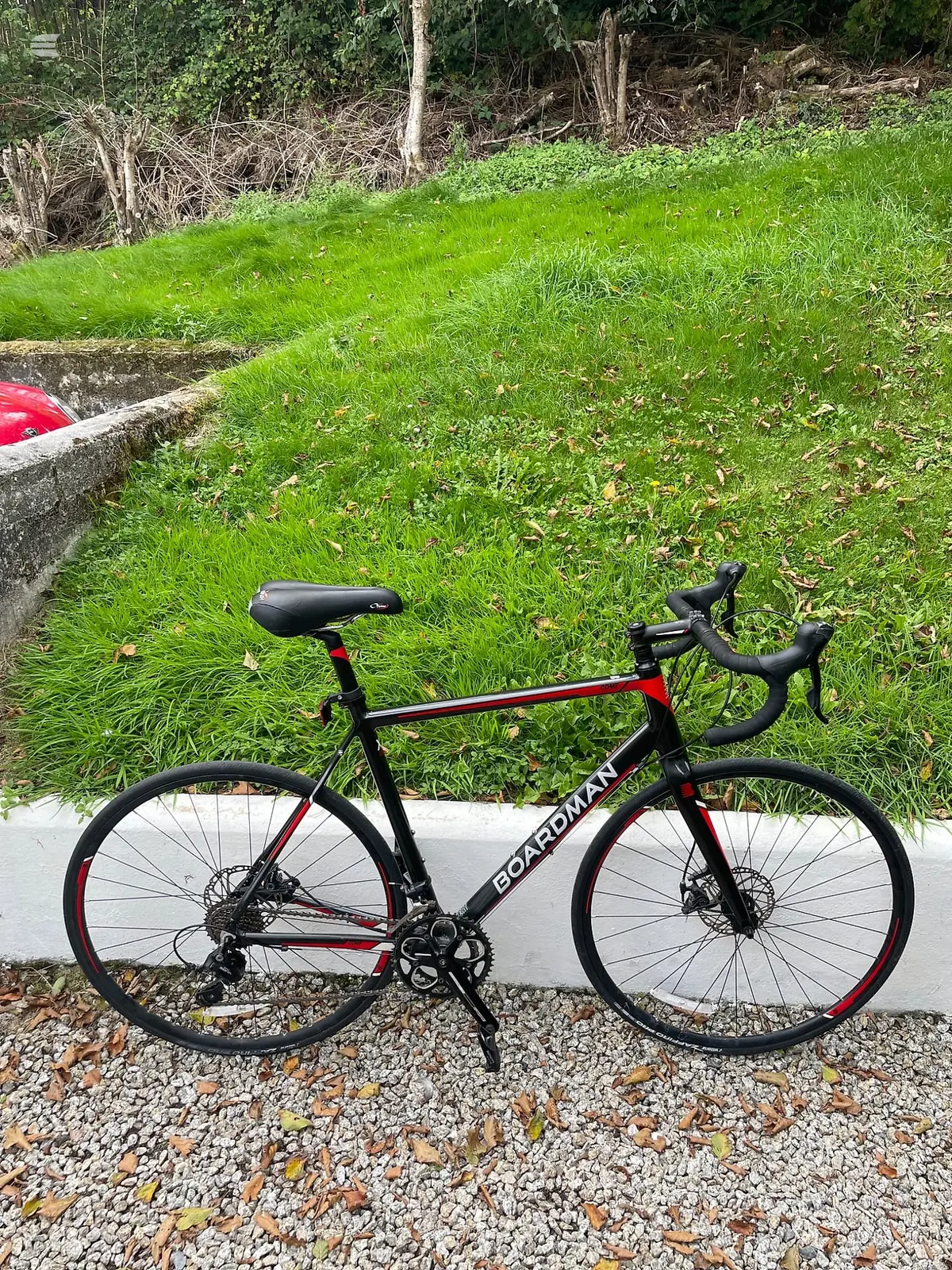 Black and red Boardman road bike with disc brakes, drive side, on gravel against a concrete curb with green lawn and woodland behind.