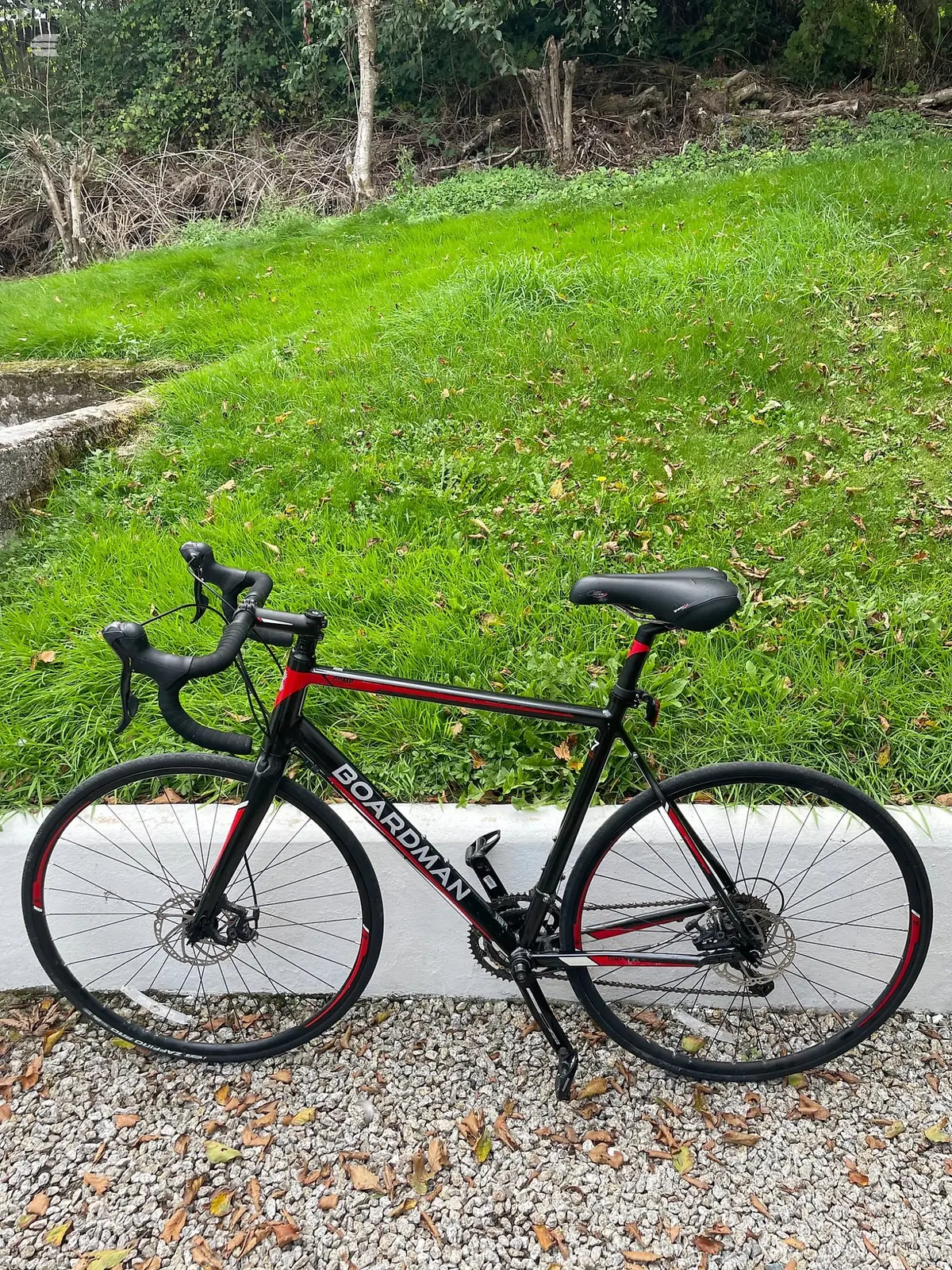 Black and red Boardman road bike with disc brakes, drive side, on gravel with grass and woodland behind.