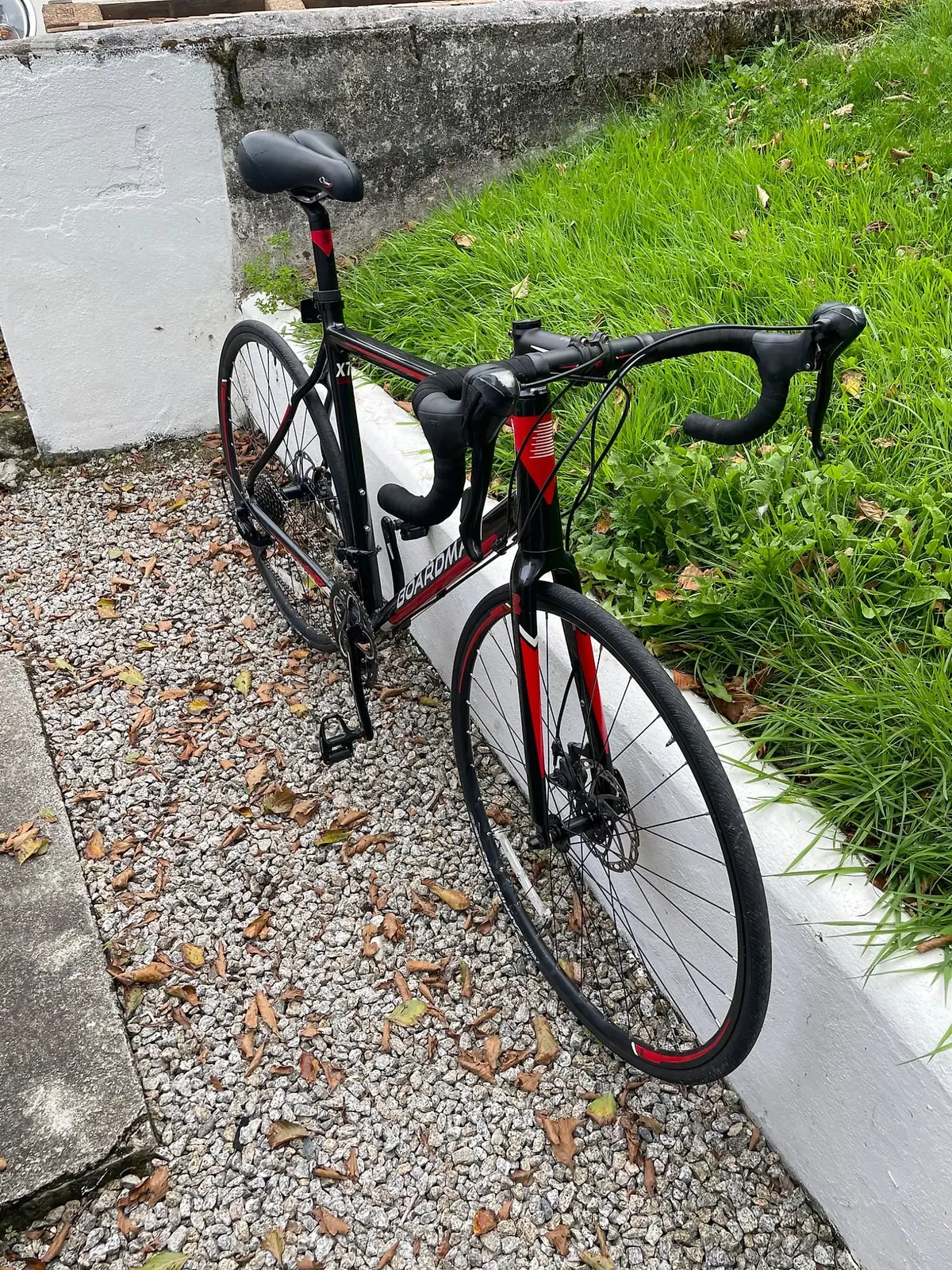Black and red Solatium road bike, drive side, leaning against a white-painted concrete wall on gravel ground with overgrown grass behind.