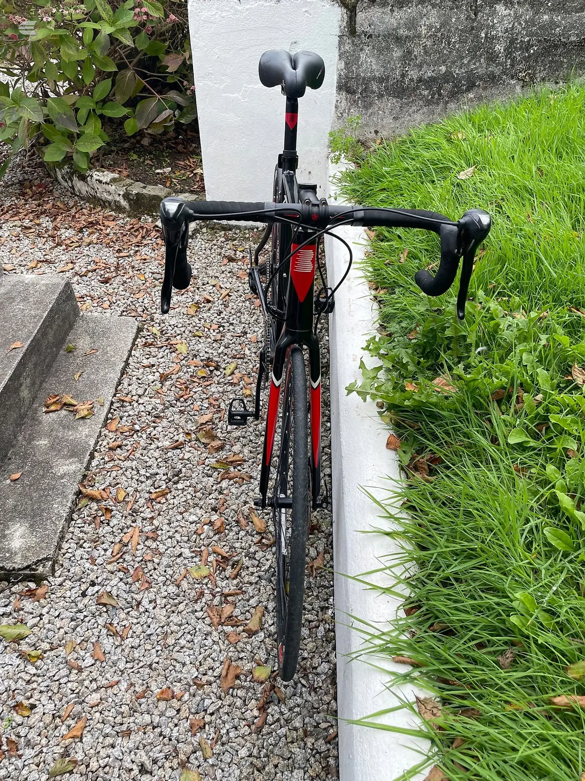 Black and red road bike, drive side, leaning against a white-painted concrete post in a residential garden setting with gravel and grass visible.