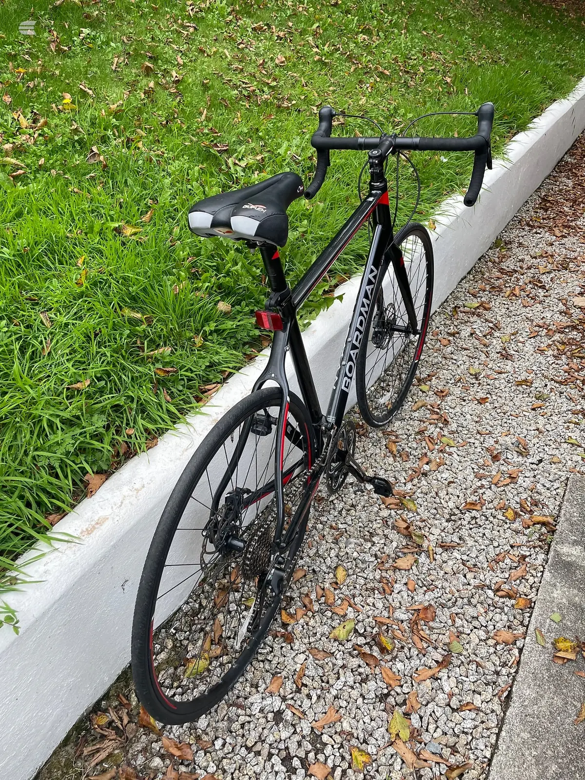 Black and red Boardman road bike, drive side, parked on gravel against a white curb with grass beyond.