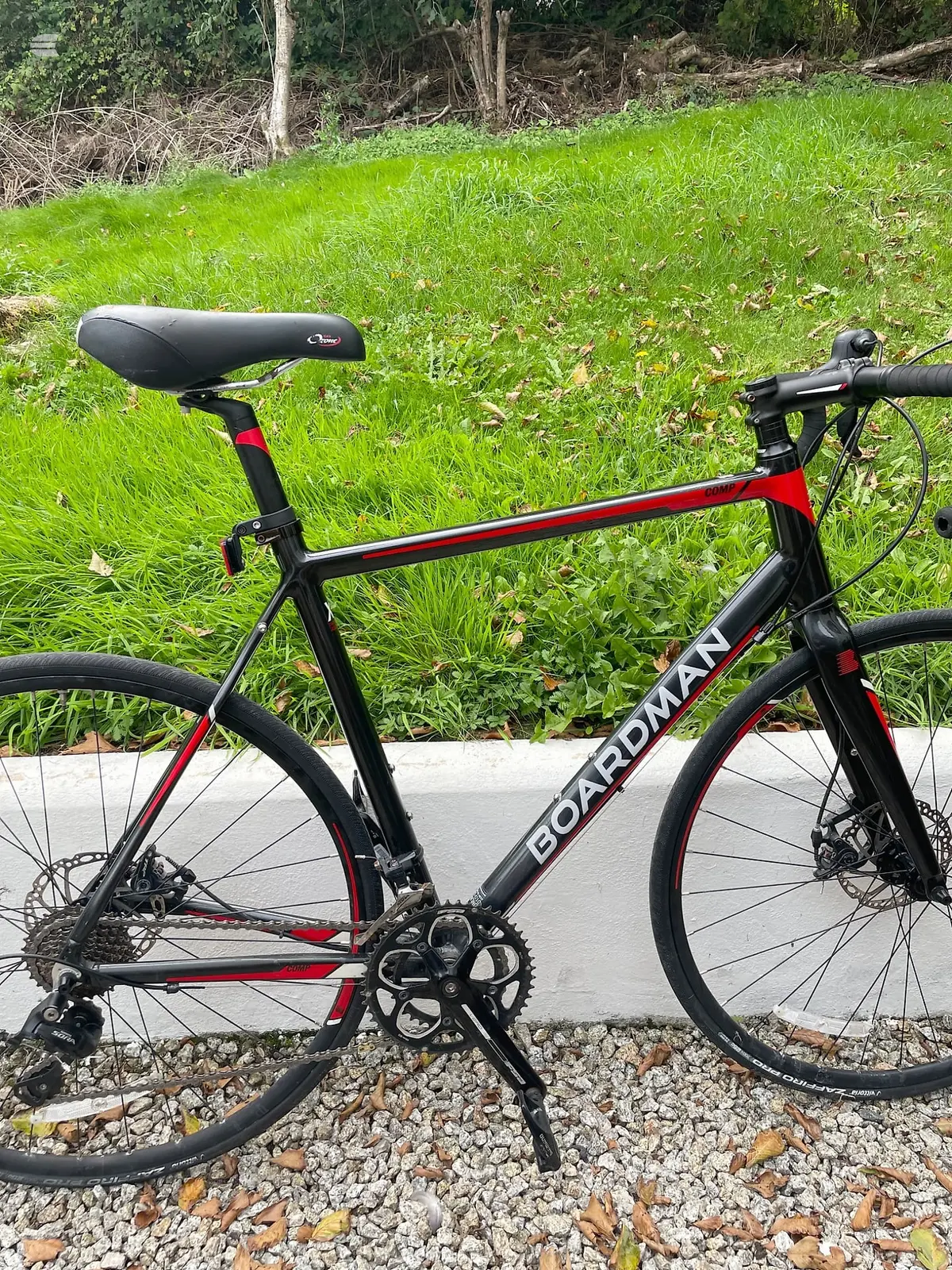 Black and red Boardman road bike on gravel, drive side, with green grass and woodland in background.