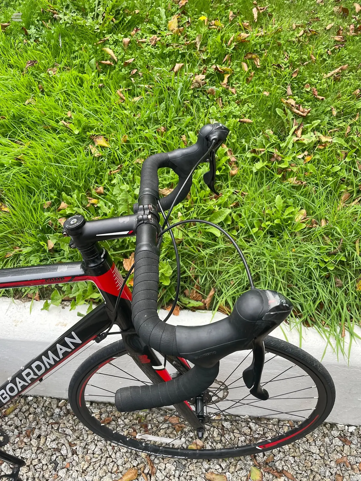 Red and black Boardman road bike on gravel, drive side view, drop bars and saddle visible, grass and fallen leaves in background.