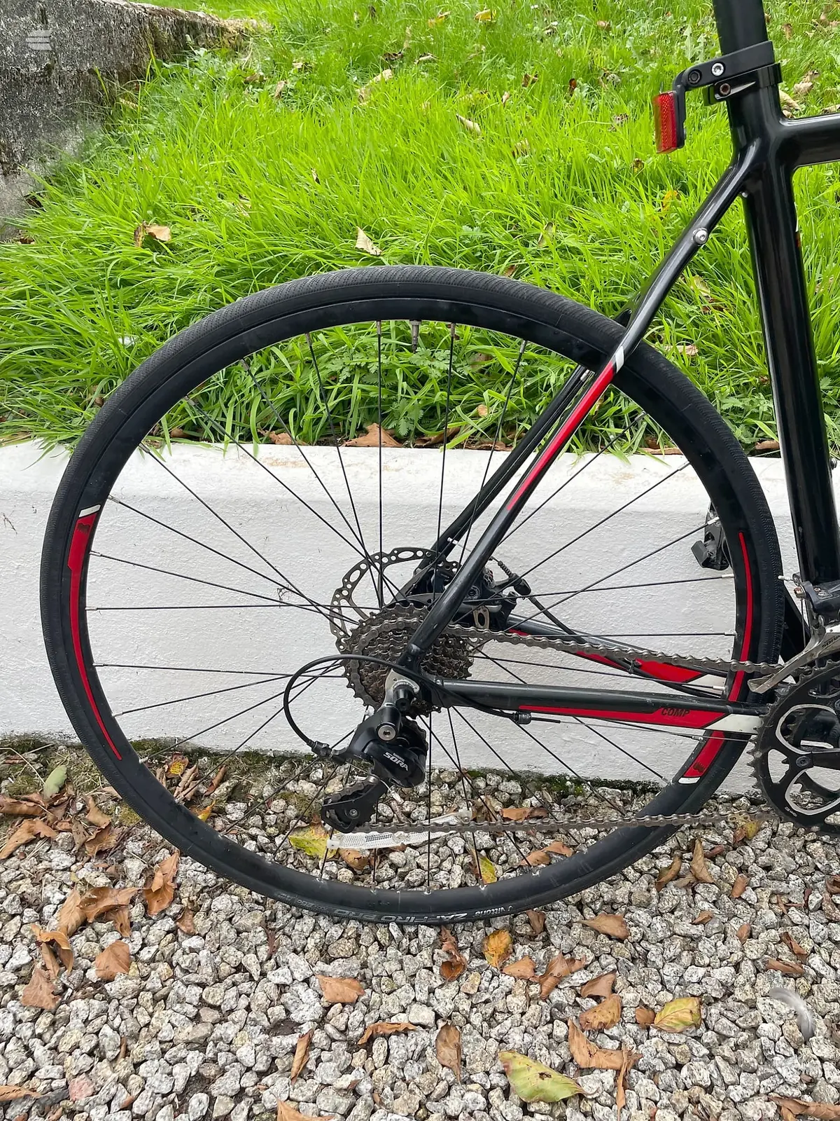 Black and red gravel bike, front wheel close-up on drive side, parked on gravel with grass and concrete in background.