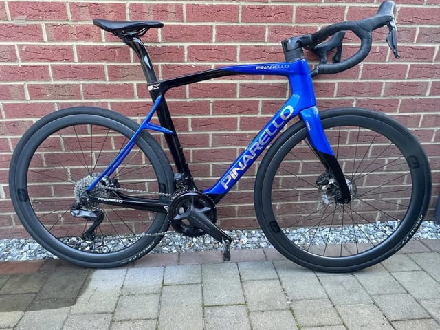 Blue and black Pinarello road bike with disc brakes, drive side, leaning against a red brick wall with white gravel at the base.