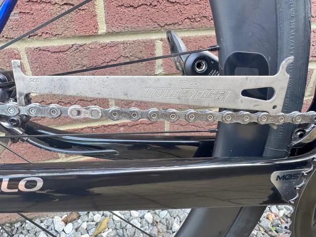 Close-up of bicycle drivetrain components: chain, derailleur, and chainring on a black frame against a brick wall background, drive side view.