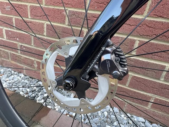 Shimano disc brake caliper and rotor on bicycle front wheel, drive side, against red brick wall with gravel ground.