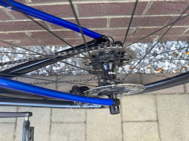 Close-up of blue road bike rear drivetrain and cassette against brick wall, drive side, showing chain, derailleur, and sprockets.