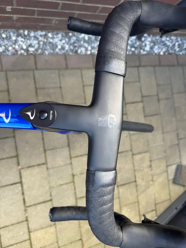 Close-up of drop bar handlebars and stem on a blue road bike, non-drive side, against a brick wall and paved ground.