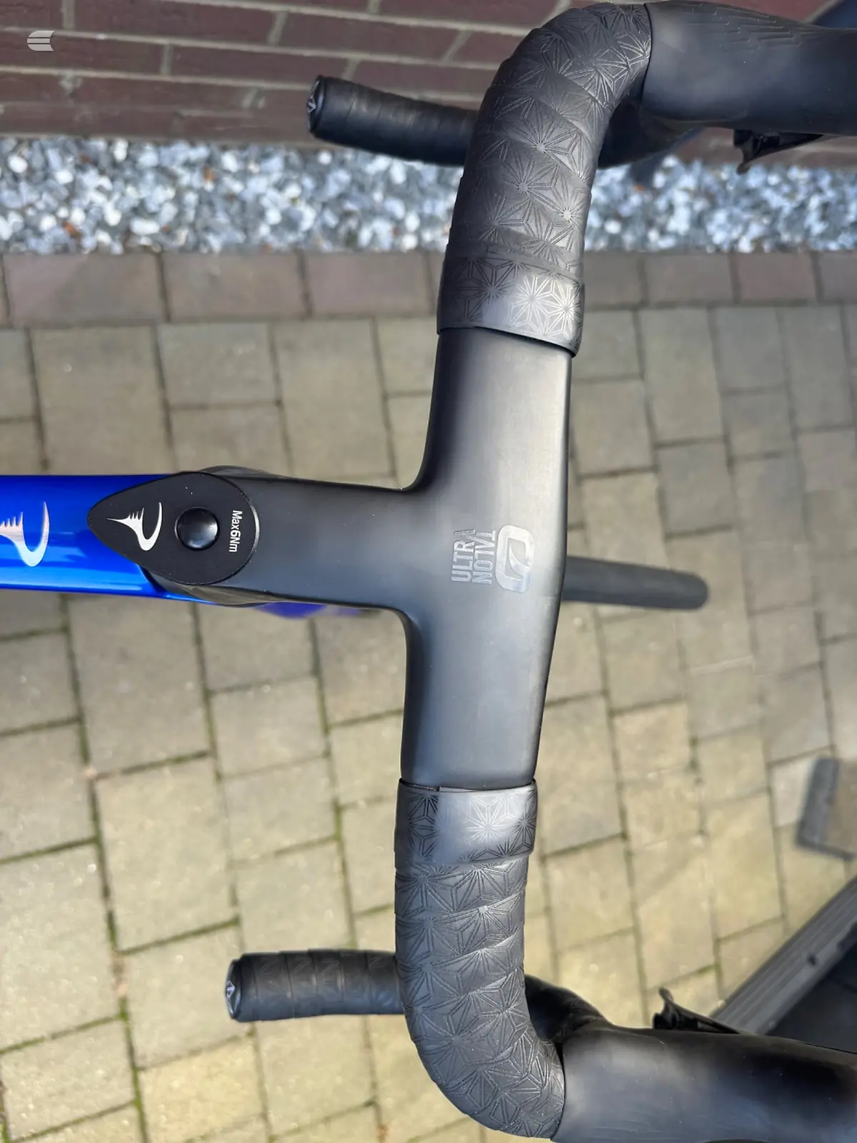 Close-up of drop bar handlebars and stem on a blue road bike, non-drive side, against a brick wall and paved ground.